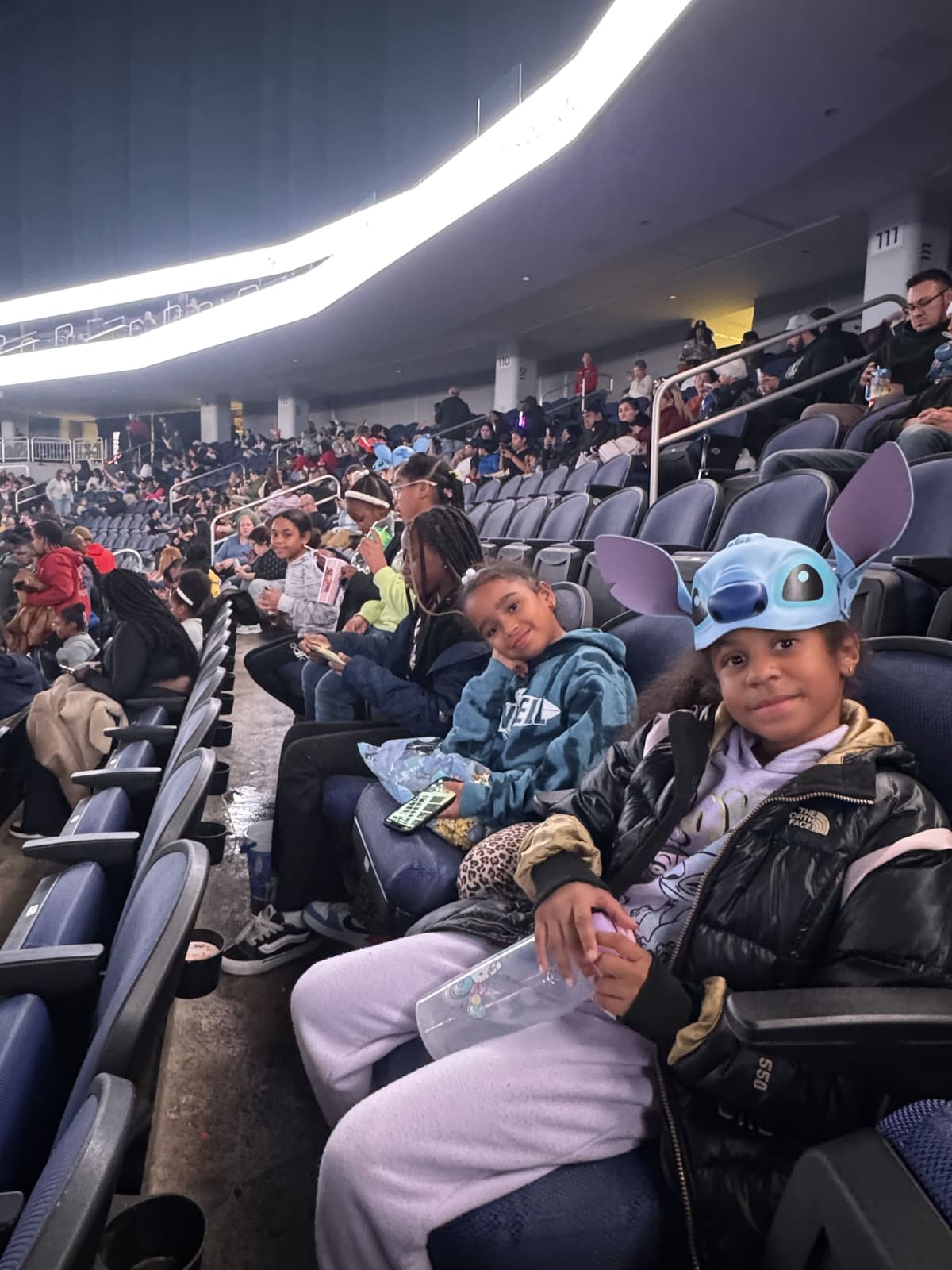CLC kids in their seats at a stadium show, one wearing a Stitch hat