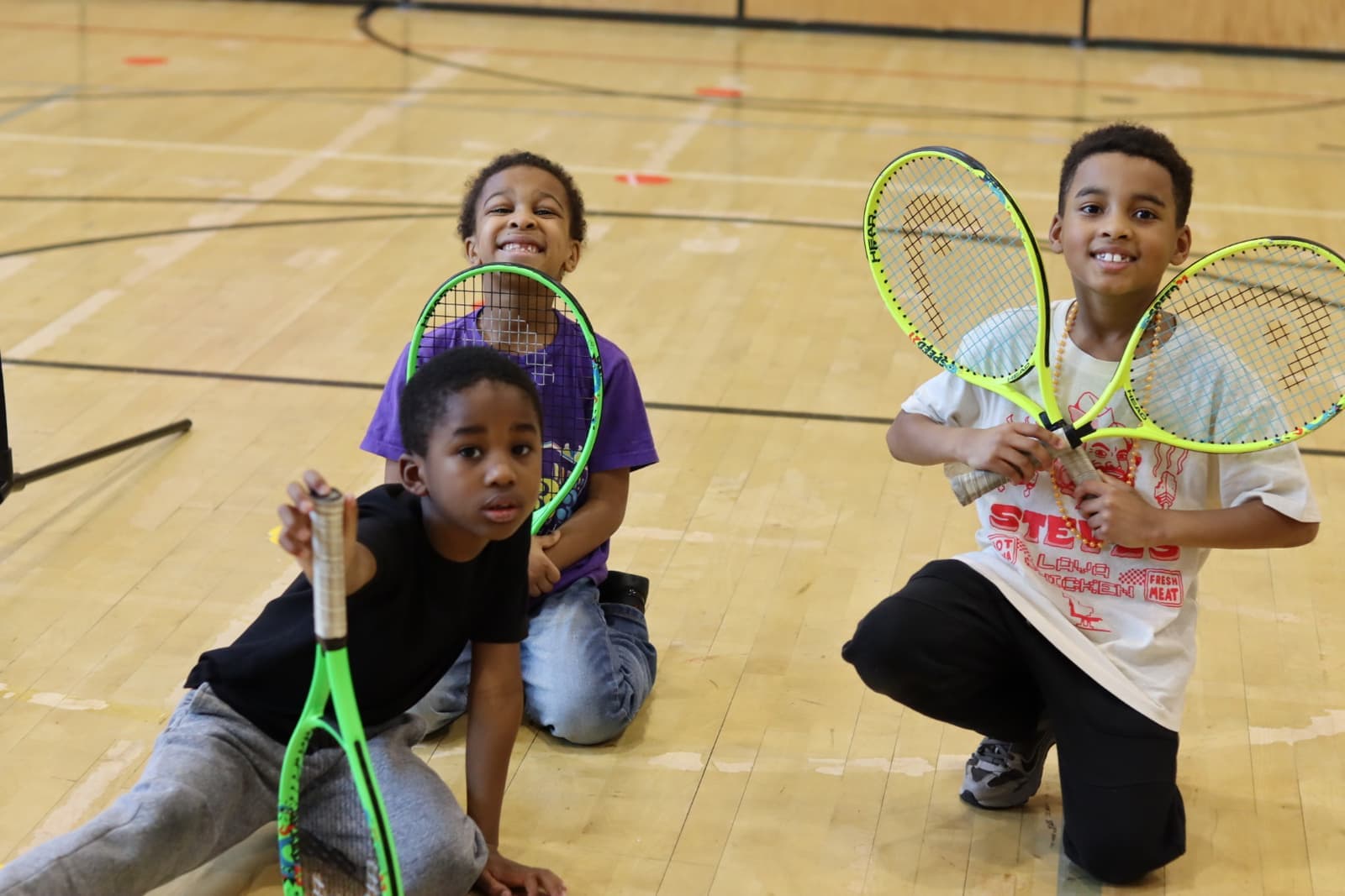 Three young boys smiling with junior rackets on the gym floor