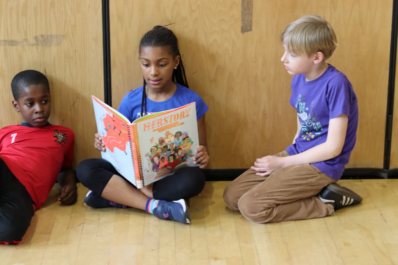 A student reading the book Herstory aloud to two classmates in the gym