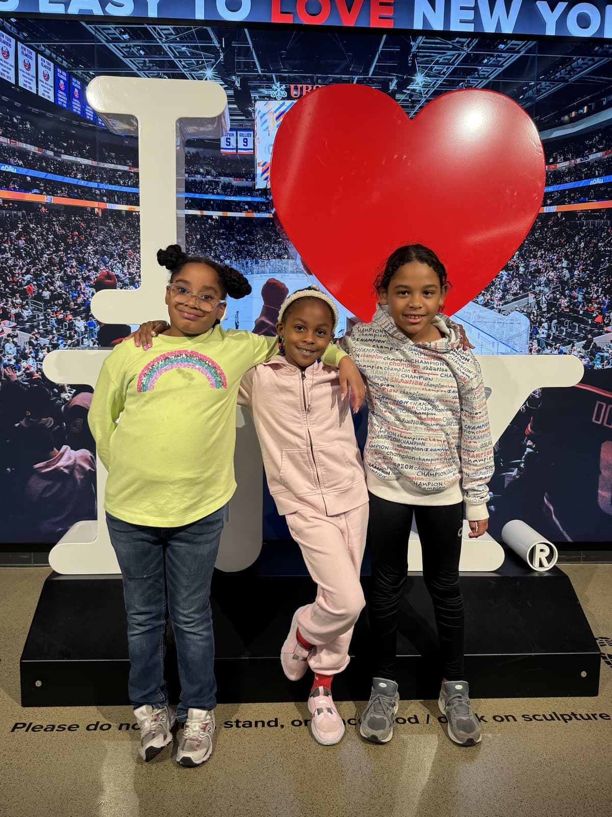 Three girls posing together in front of an 'I love New York' arena display