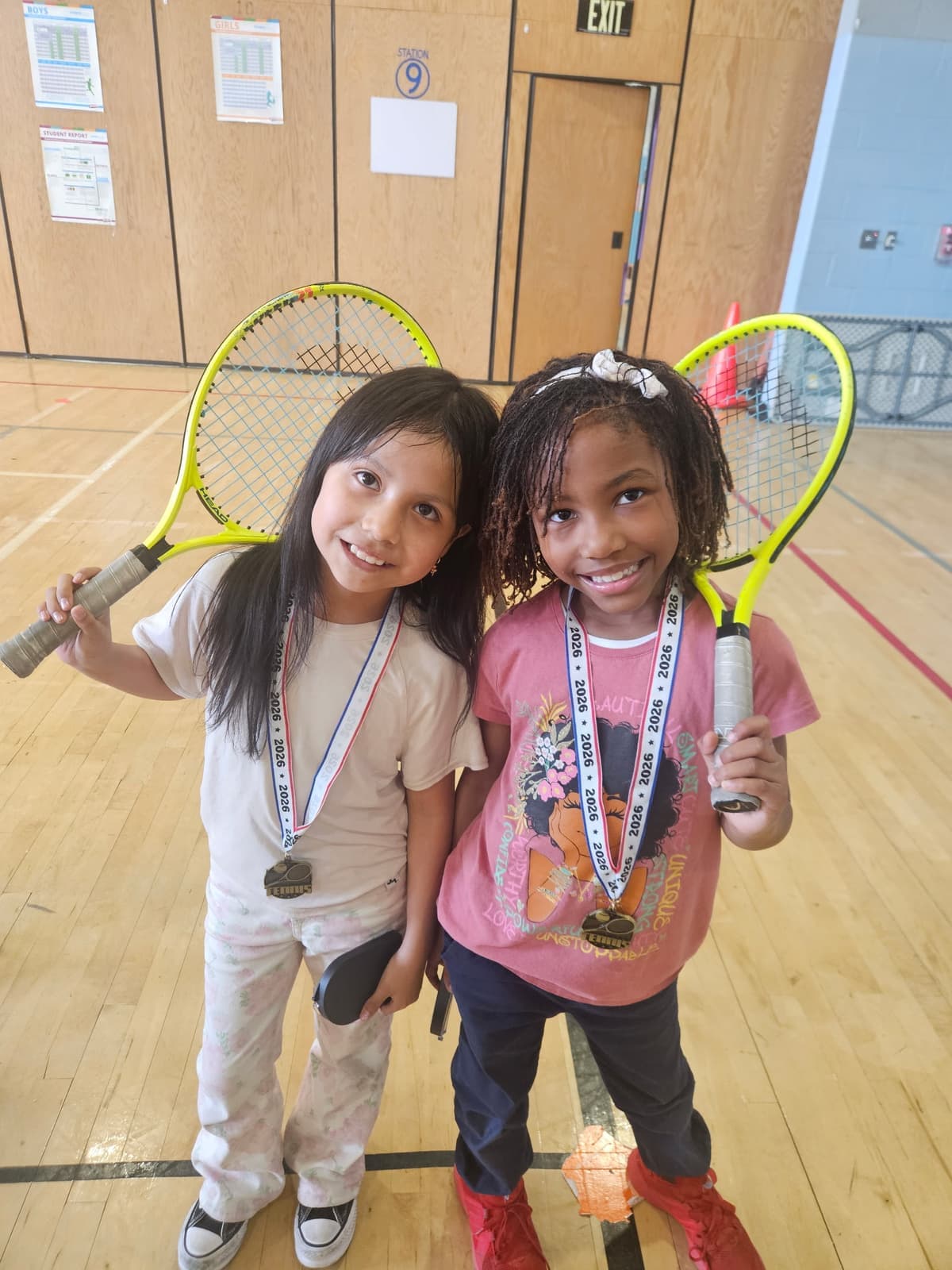 Two girls wearing 2026 medals and holding rackets overhead